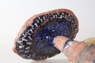 Nahaufnahme der Unterseite einer Keramikskulptur, die einem Pilz ähnelt, mit einem strukturierten Stiel und einem dunkelblauen, gesprenkelten Hut mit weißen, gelben und schwarzen Details.