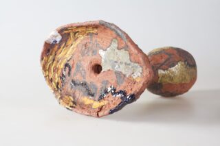 Eine strukturierte, abstrakte Keramikskulptur mit einem breiten, ovalen Ende mit bunten bemalten und metallischen Flecken, die auf der Seite vor einem schlichten Hintergrund liegt.