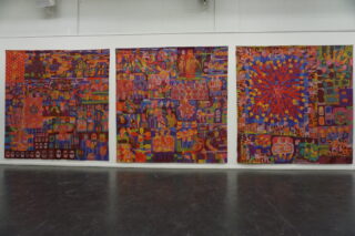 Drei große, farbenfrohe, abstrakte Gemälde mit komplizierten Mustern und Formen hängen nebeneinander an einer weißen Wand in einem Galerieraum.