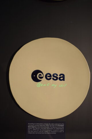 Ein beiges, rundes Schild zeigt ein modifiziertes ESA-Logo mit dem Schriftzug „Eat my shit“ in blauer und grüner Schrift. Unter dem Schild ist ein Etikett im Museumsstil zu sehen.