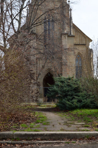 Eine Steinkirche mit gotischen Bögen und hohen Fenstern wird teilweise von blattlosen Bäumen und überwucherten Sträuchern vor einem ausgetretenen Pfad verdeckt.