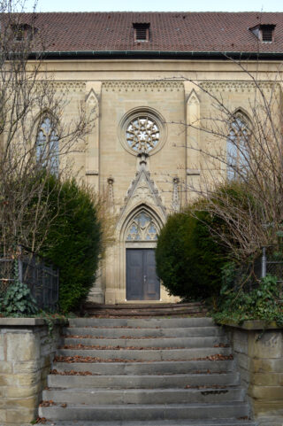 Steinstufen führen zu einer geschlossenen Holztür eines Gebäudes im gotischen Stil mit Bogenfenstern, einem runden Buntglasfenster und überwucherten Büschen auf beiden Seiten.