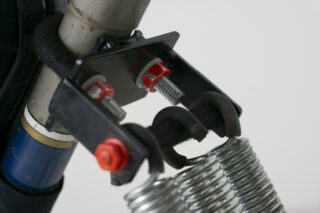 Nahaufnahme einer mechanischen Baugruppe mit Metallfedern, Bolzen und einer schwarzen Halterung, die an einer zylindrischen blau-silbernen Struktur befestigt ist.