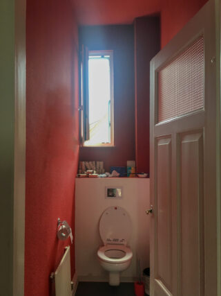 Ein kleines Badezimmer mit roten Wänden, einer weißen Toilette, einem offenen Fenster über der Toilette und verschiedenen Toilettenartikeln auf dem Regal dahinter.
