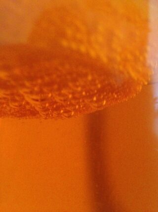 Nahaufnahme einer orangefarbenen Flüssigkeit mit Blasen nahe der Oberfläche, die ein strukturiertes und abstraktes Erscheinungsbild erzeugen.