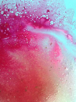 Nahaufnahme einer rosa und weißen Flüssigkeit mit Blasen und strukturierten Mustern, die eine Mischung oder Reaktion auf einer glatten Oberfläche suggerieren.