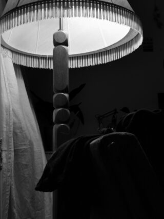 In einem dunklen Raum brennt eine Stehlampe mit einem Schirm mit Fransen, neben einem drapierten Tuch und einem Stuhl mit dunklem Stoff.