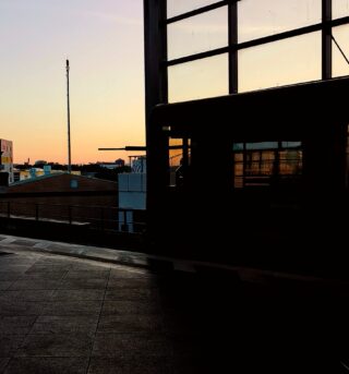 Ein Busbahnhof bei Sonnenuntergang mit einem Bus im Schatten, großen Fenstern und im Hintergrund sichtbaren Gebäuden.