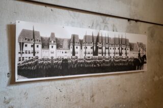 Ein Schwarzweißfoto einer Gruppe uniformierter Soldaten, die in Formation vor einem großen Gebäude stehen, montiert auf einer Betonmauer.