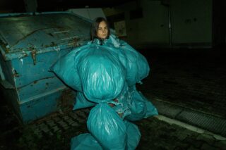 Eine in blaue Müllsäcke gekleidete Person steht nachts neben einem großen Müllcontainer in einer städtischen Gasse.