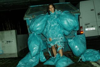 Eine Person steht nachts vor einem Müllcontainer. Sie trägt ein Kleid und übergroße Ärmel aus blauen Plastikmüllsäcken.