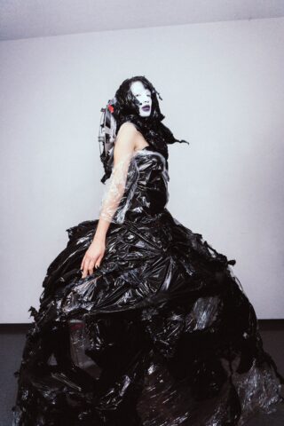 Eine Person trägt ein dramatisches schwarzes Kleid aus Kunststoffmaterialien und transparenten Elementen, mit weißem Make-up und schwarzem Lippenstift und steht vor einem schlichten Hintergrund.