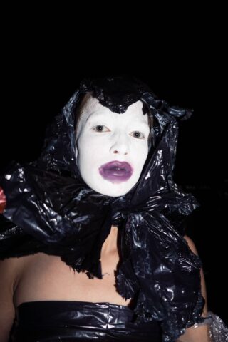 Person mit weißer Gesichtsbemalung und dunklem Lippenstift, die ein schwarzes Plastikmaterial als Kapuze und Kleidungsstück trägt und vor einem dunklen Hintergrund posiert.