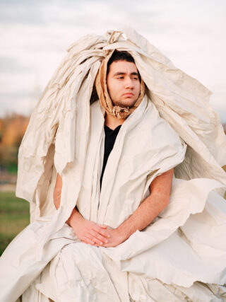 Eine Person, die eine große Menge zerknülltes beiges Papier über Kopf und Körper trägt, steht mit gefalteten Händen im Freien.
