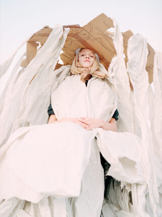 Vor einem hellen Hintergrund sitzt eine Person mit gefalteten Händen, die in weißen Stoff gehüllt ist und eine Pappkonstruktion als Hut trägt.