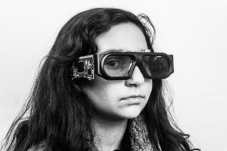 Eine junge Person mit langen Haaren trägt eine große, experimentelle elektronische Brille mit sichtbaren Leiterplatten und blickt vor einem schlichten Hintergrund direkt in die Kamera.