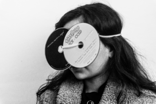 Dabei trägt eine Person zwei CDs wie eine Schutzbrille vor den Augen, die mit einem Gummiband um den Kopf befestigt sind.