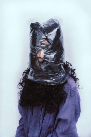 Eine Person mit langem, lockigem Haar und einem lila Hemd trägt eine durchsichtige Plastiktüte, die Kopf und Gesicht bedeckt.