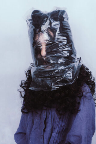 Eine Person mit langen lockigen Haaren trägt eine durchsichtige Plastiktüte über Kopf und Schultern, ist mit einem weiten lila Hemd bekleidet und steht vor einem blassen Hintergrund.