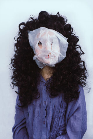 Eine Person mit langem, lockigem, dunklem Haar trägt ein lockeres, halbtransparentes weißes Laken über dem Gesicht und ein lila Hemd vor einem schlichten hellen Hintergrund.