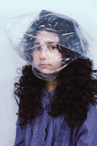 Eine Person mit langem lockigem Haar und einem lila Hemd trägt vor einem schlichten Hintergrund eine durchsichtige Plastiktüte auf dem Kopf.