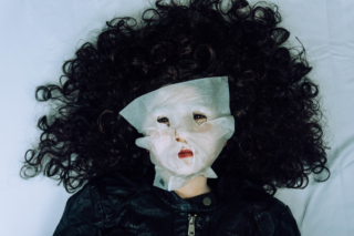 Eine Puppe mit lockigem dunklem Haar und einer schwarzen Jacke liegt auf einer weißen Fläche, ihr Gesicht ist von einer weißen Gesichtsmaske bedeckt.
