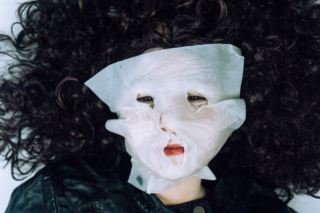 Eine Person mit lockigem dunklem Haar trägt eine weiße Gesichtsmaske, die ihr Gesicht teilweise bedeckt, liegt da und ist mit einer dunklen Jacke bekleidet.