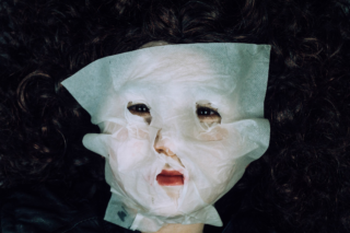 Eine Nahaufnahme einer Person, die eine weiße Gesichtsmaske trägt, mit sichtbarem lockigem Haar und teilweise freiliegenden Lippen und Augen.