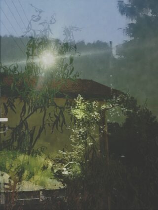 Ein verschwommenes, künstlerisches Foto zeigt ein Haus mit rotem Dach, Bäumen und Sonnenlicht, das durch Glas reflektiert wird oder scheint, wodurch vielschichtige, abstrakte Reflexionen entstehen.