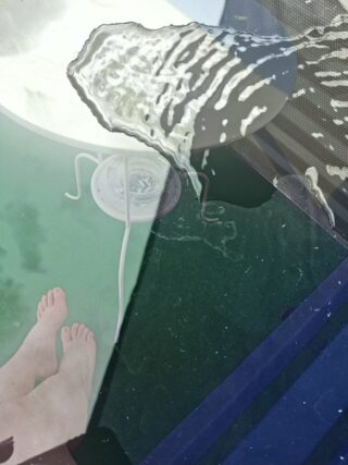 Ein Paar nackte Füße neben einem Billardtisch, über dessen Rand Wasser läuft, das Spiegelbilder und Schatten auf die nasse Oberfläche wirft.