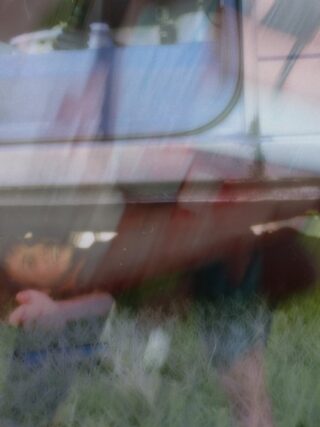 Ein verschwommenes, doppelt belichtetes Bild, das eine Person zeigt, die im Gras unter einem Fahrzeug liegt, wobei eine undeutliche Mischung aus Farben und Formen die Szene überlagert.