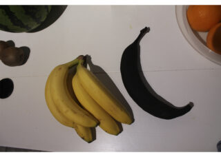 Vier gelbe Bananen und eine überreife schwarze Banane liegen auf einer weißen Fläche neben einer Wassermelone, Kiwis und einer Schale Orangen.