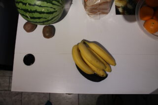 Eine Bananenstaude auf einem weißen Tisch, daneben eine Wassermelone, zwei Kiwis, ein Laib Brot, eine Schale Orangen und ein dunkles rundes Objekt.