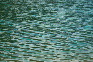 Nahaufnahme von plätscherndem Wasser auf einer Wasserfläche. Die Oberfläche reflektiert Licht und erscheint grünlich-blau.