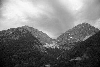 Schwarzweißfoto von zwei felsigen, bewaldeten Bergen unter einem bewölkten Himmel. An den Hängen sind kleine Schneeflecken sichtbar.