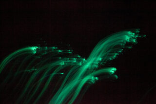 Grüne Lichtspuren krümmen sich und verschwimmen vor einem schwarzen Hintergrund, wodurch ein abstrakter Bewegungseffekt entsteht.