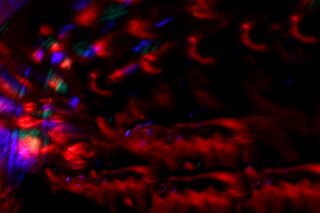 Abstraktes Bild mit wirbelnden roten, blauen und grünen Lichtmustern auf dunklem Hintergrund, wobei im unteren Bereich ein horizontales Gitter sichtbar ist.