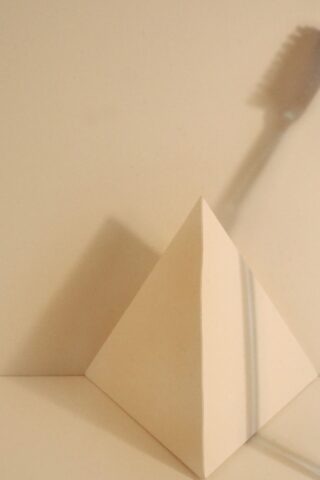 Eine beige Pyramide steht auf einer flachen Oberfläche vor einem passenden Hintergrund, an die Wand wirft sich der Schatten einer Zahnbürste.
