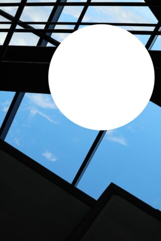 Blick auf eine Glasdecke mit sichtbarem blauen Himmel und Wolken, unterbrochen von einem schwarzen Rahmen und einem großen weißen kreisförmigen Bereich in der Mitte.