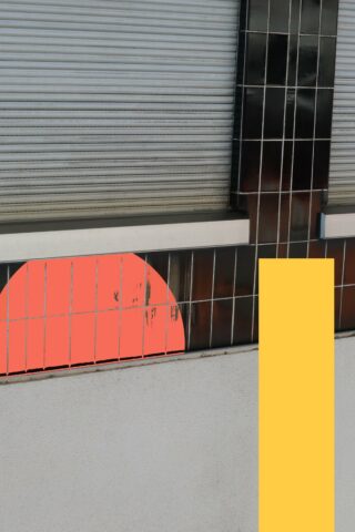 Eine Wand mit schwarzen Fliesen, einem teilweise sichtbaren orangefarbenen Halbkreis, einem vertikalen gelben Rechteck und einem Metallladen darüber.