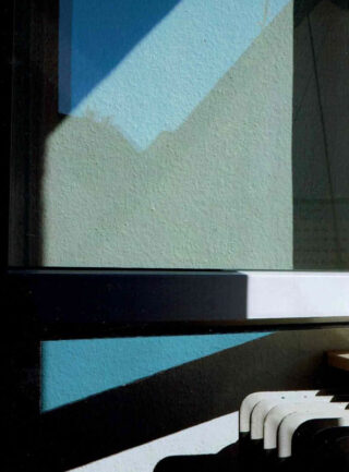 Geometrische Schatten und Lichtmuster fallen über eine Wand, ein Fenster und einen Teil einer weißen Bank.