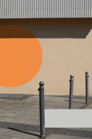 Drei graue Poller stehen auf einem gepflasterten Gehweg neben einer beigen Mauer, die einen orangefarbenen Halbkreis und einen Schatten werfenden Überhang aus Wellblech aufweist.