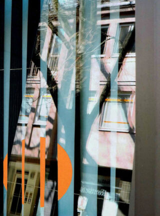 Spiegelung eines Gebäudes und eines Baums in einem Glasfenster mit vertikalen blauen Streifen und orangefarbenen geometrischen Formen im Vordergrund.