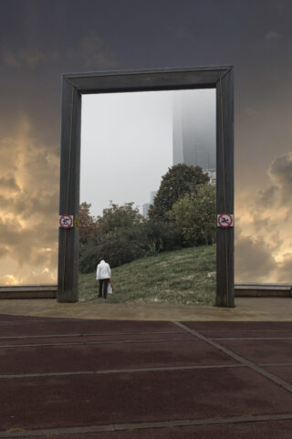 Eine Person im weißen Kittel geht durch einen großen rechteckigen Rahmen im Freien. Dahinter ist ein bewölkter Himmel zu sehen, im Inneren ist Grün zu sehen. Auf dem Rahmen sind zwei Schilder mit der Aufschrift „Hunde verboten“ angebracht.