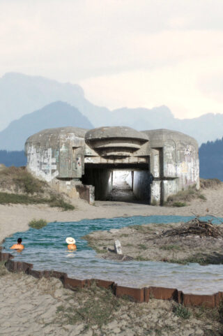 Zwei Personen sitzen im seichten Wasser in der Nähe eines verwitterten, mit Graffiti bedeckten Betonbunkers, der in einer Sandlandschaft mit fernen Bergen im Hintergrund liegt.