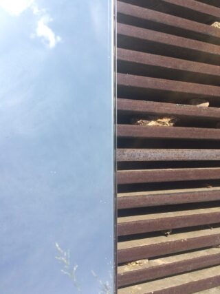 Eine vertikal geteilte Ansicht, die links eine reflektierende, hellblaue Oberfläche und rechts ein rostiges Metallgitter mit horizontalen Lamellen und Schutt zeigt.