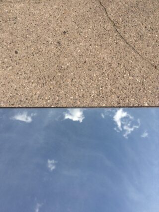 Über einem Spiegel ist eine Betonoberfläche zu sehen, die einen blauen Himmel mit vereinzelten Wolken reflektiert.