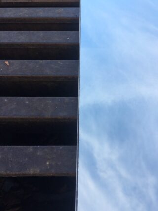 Eine Nahaufnahme eines Metallgitters auf der linken Seite neben einer glatten Oberfläche, die einen blauen Himmel mit Wolken auf der rechten Seite reflektiert.