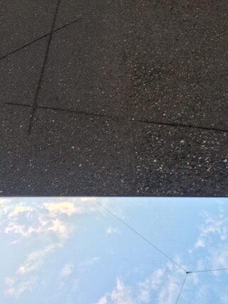 Eine reflektierende Oberfläche auf dem Boden spiegelt den Himmel mit Wolken wider, während darüber ein strukturierter Bürgersteig mit sich kreuzenden Linien sichtbar ist.
