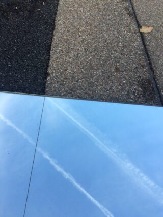 Eine reflektierende Oberfläche zeigt blauen Himmel und Kondensstreifen, die neben strukturiertem Kies und Asphaltbelag liegen.
